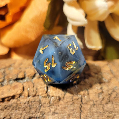 A twenty-sided die that has a clear base with black, white, and Oxford blue drips. The numbers are inked in gold, and the most prominent ones from this angle are 1, 7, 13, 15, 5, and 12. The die colors from this angle look like a milky blue swirl, and gold flakes are reflected in the lighting.