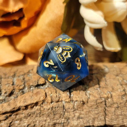 A twenty-sided die that has a clear base with black, white, and Oxford blue drips. The numbers are inked in gold, and the twenty-face is the logo of a bee. The prominent numbers displayed are 2, 14, 8, 12, 10, and 20 as the logo.
