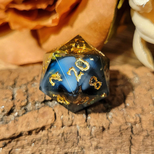 A twenty-sided die that has a clear base with black, white, and Oxford blue drips. The font is Sugar Bee RPG's signature death metal font, and is inked in gold. The most prominent numbers are 14, 20, 2, and 8. Large gold foil flakes are suspended within the die.