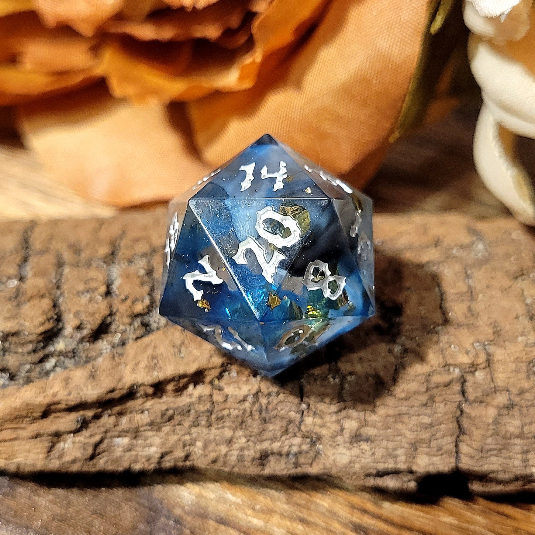 A twenty-sided die that has a clear base with black, white, and Oxford blue drips. The font is Sugar Bee RPG's signature death metal font, and is inked in white. The most prominent numbers are 20, 14, 2, and 8. Large gold foil flakes are suspended within the die. This angle is a beautiful bright blue with gold flake accents.