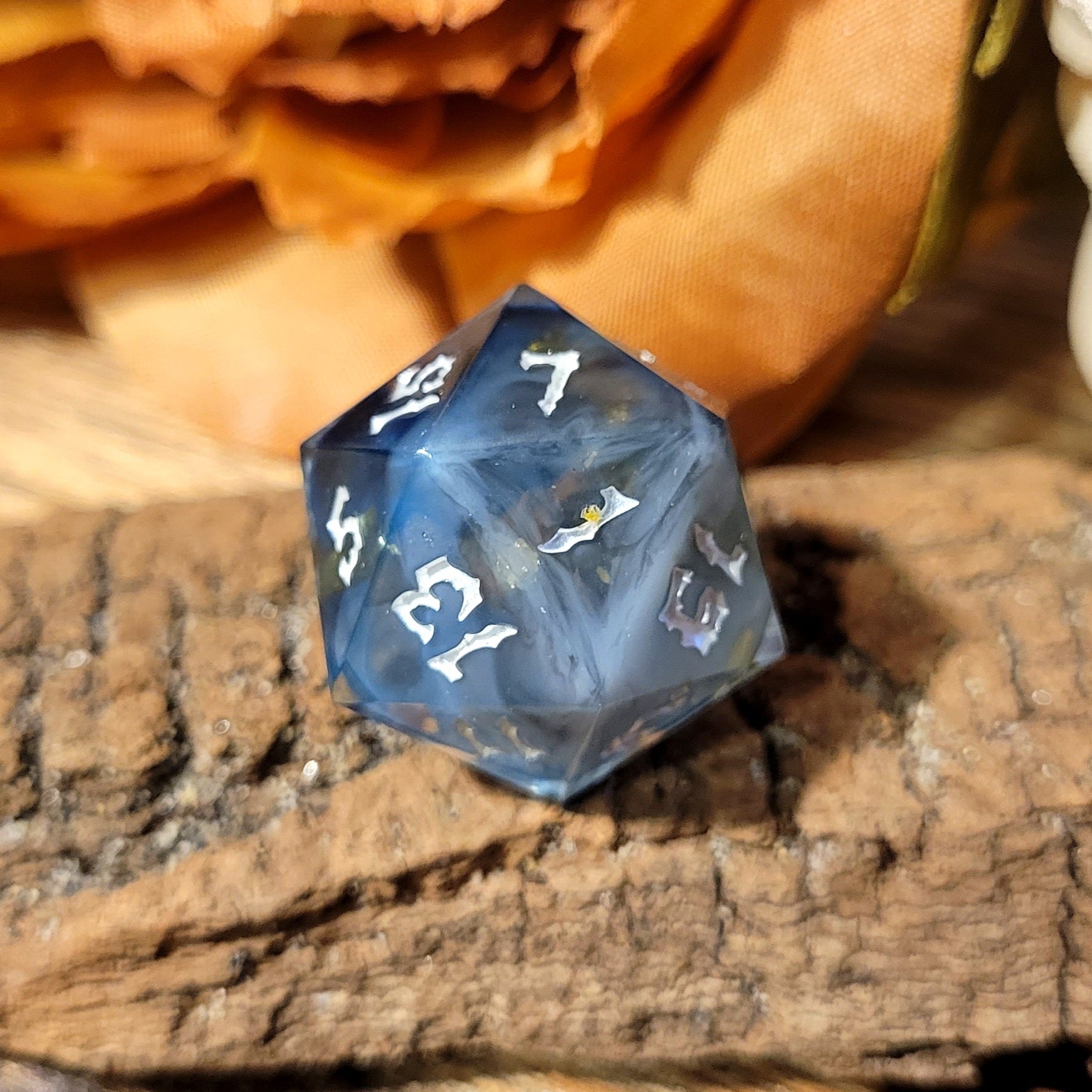 A twenty-sided die that has a clear base with black, white, and Oxford blue drips. The font is Sugar Bee RPG's signature death metal font, and is inked in white. The most prominent numbers are 5, 15, 7, 13, 1 and 19. This angle has a light blue milky swirl to it.
